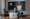 Homem sentado em frente a um notebook MacBook, com imagens realistas de uma mulher criada por inteligência artificial exibidas em um monitor grande