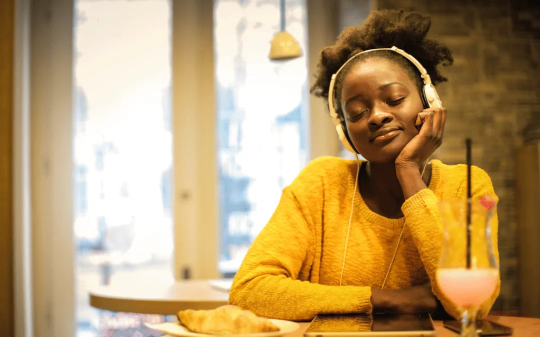 Mulher com fones de ouvido brancos, de olhos fechados e expressão relaxada, sentada em uma cafeteria com tablet e bebida ao lado.