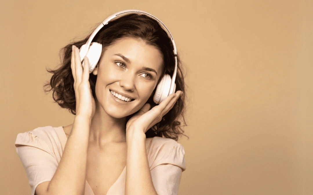 Mulher sorridente usando fones de ouvido brancos, apreciando música com fundo neutro em tom bege.