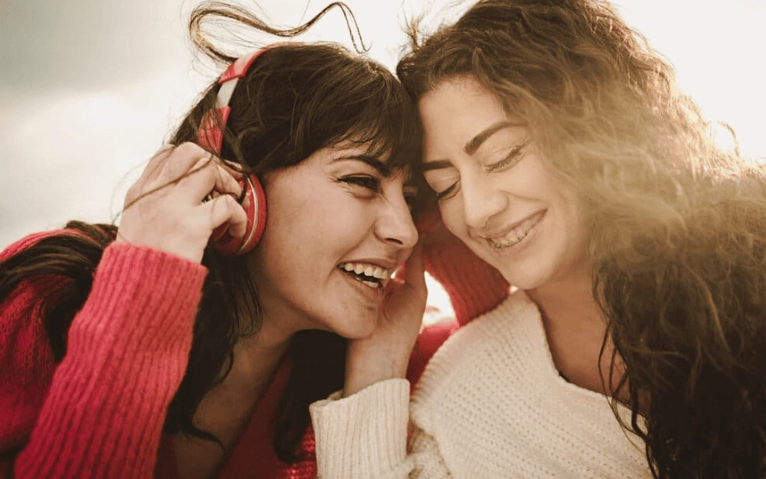 Duas amigas sorrindo e compartilhando música em fones de ouvido vermelhos ao ar livre, sob a luz do sol.