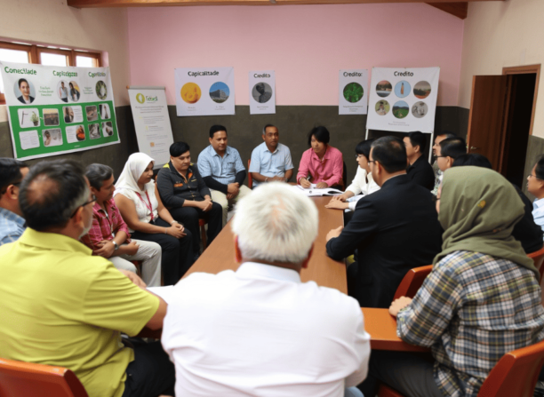 Grupo diverso de pessoas reunidas em círculo em uma sala simples, com cartazes sobre conectividade, capacitação e crédito nas paredes, representando uma reunião sobre políticas públicas no meio rural.